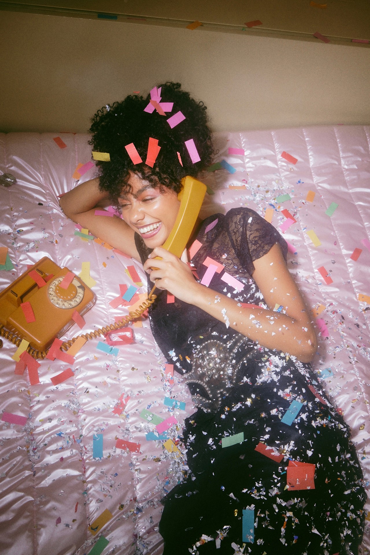 A person lies on a pink surface, wearing a colorful outfit, with hair accessories, appearing relaxed and contemplative.