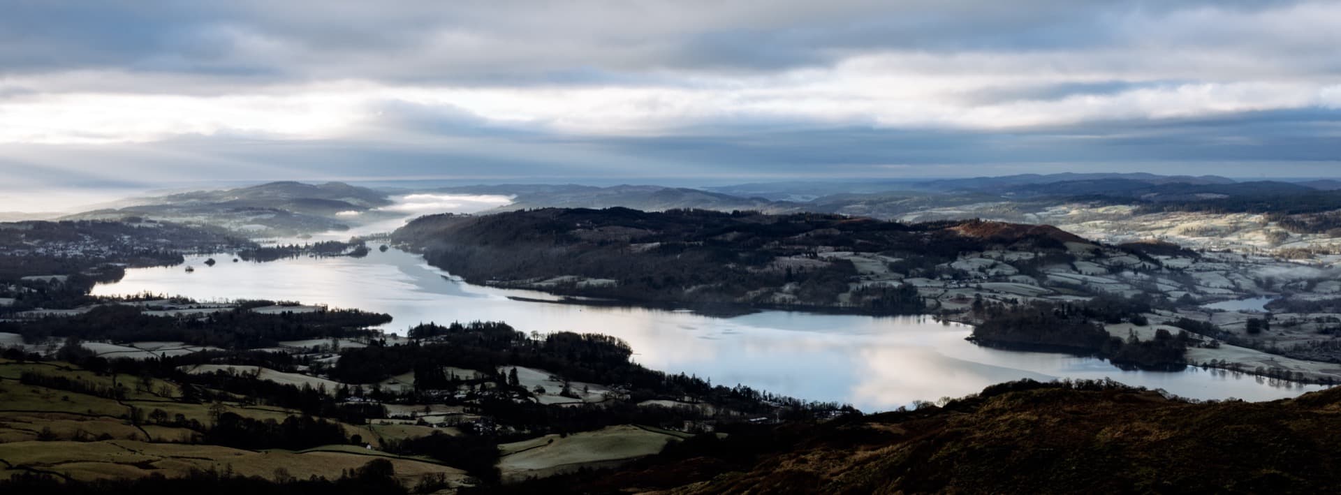 Panoramic view from Wansfell Pike across Windermere lake winding through valley floor, forested wooded islands and peninsulas, frost-covered fields and scattered settlements below winter sky