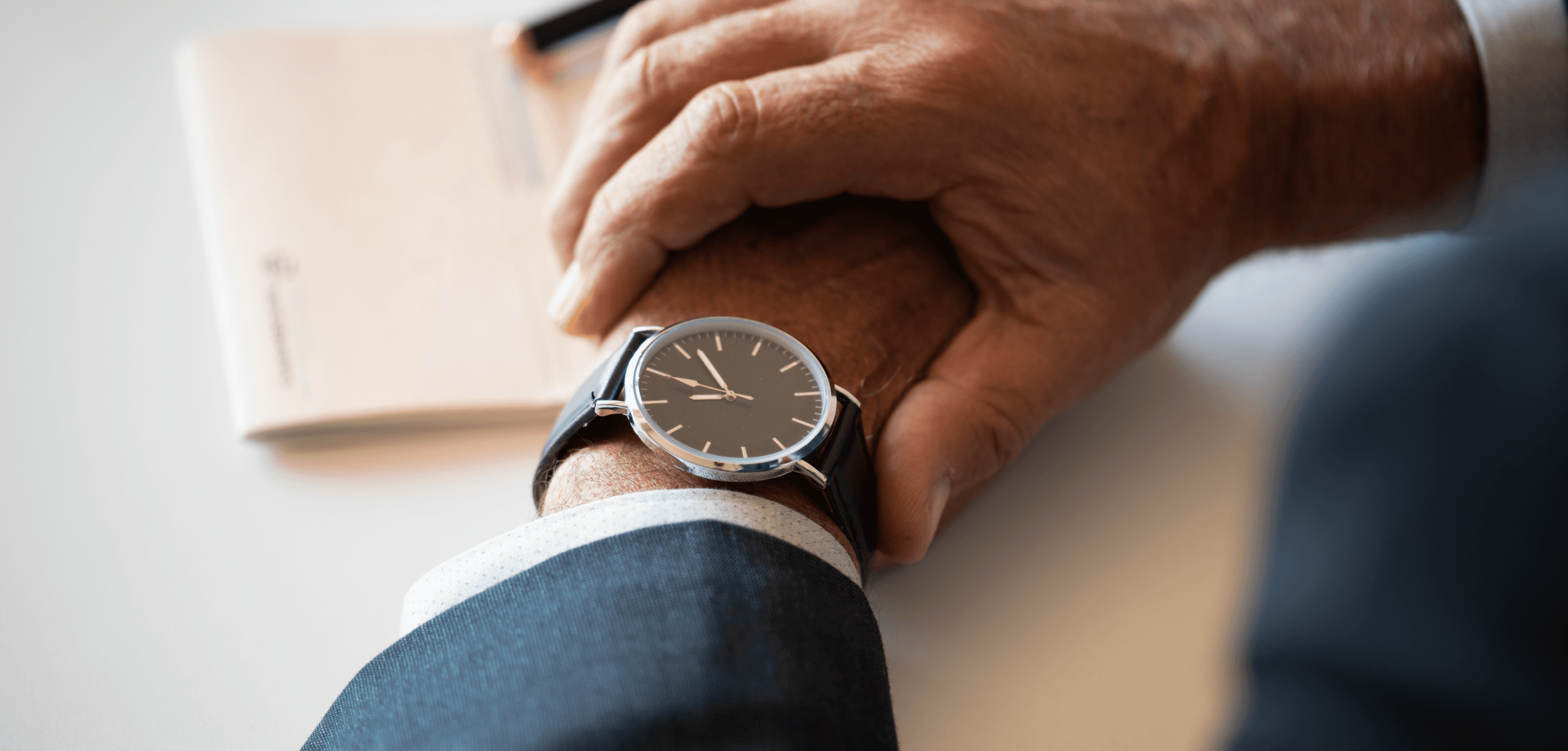 Close-up of a man in a suit checking a wristwatch with a black leather strap, with a notebook blurred in the background.