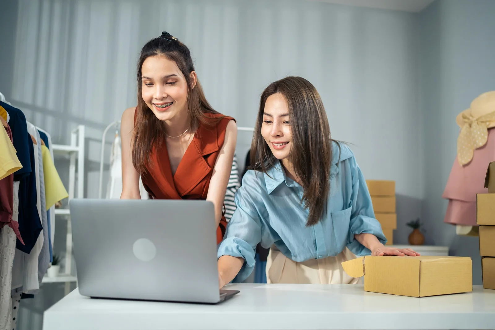 Two women smiling while reviewing laptop and cardboard boxes in a bright workspace with clothing racks.