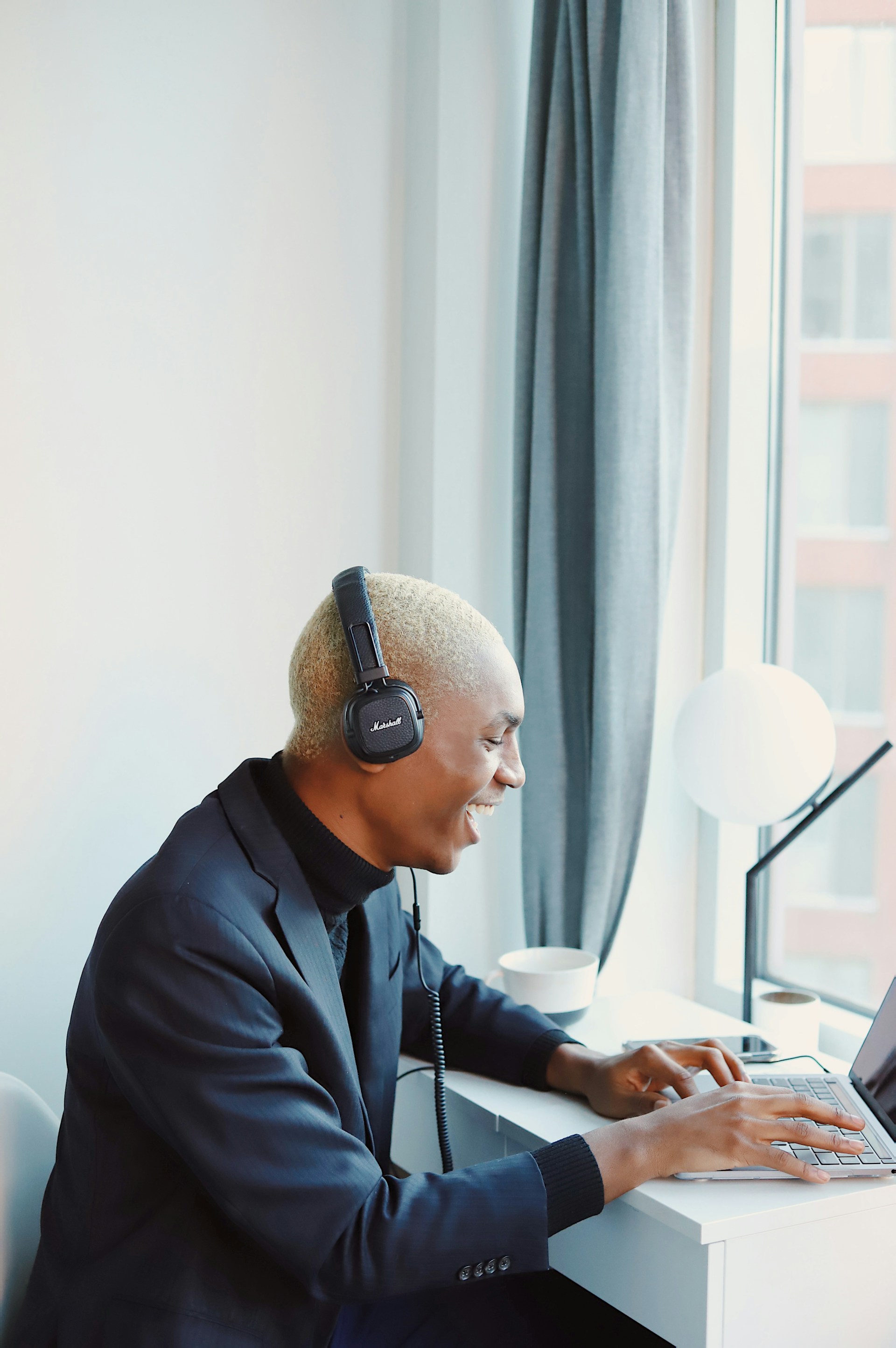 A man with buzzed blonde hair wearing a dark blazer over a black turtleneck and black Marshall headphones. He is sitting at a white desk by a window, laughing while typing on a laptop.
