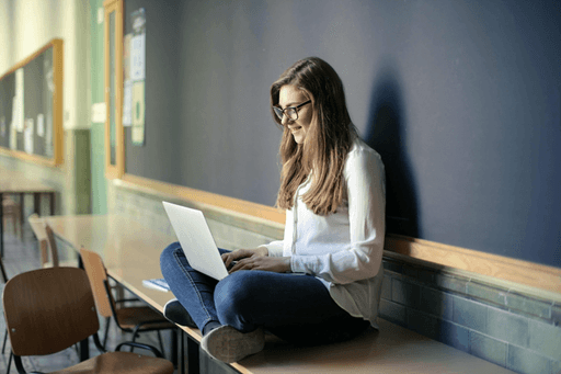 A person sitting on a bench with a laptop, focused and working in a bright, classroom-like environment.