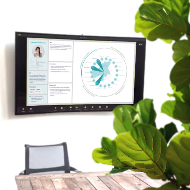 A wall-mounted screen displays a circular data chart and profile information next to a wooden table, empty chair, and green leafy plant in a meeting room.