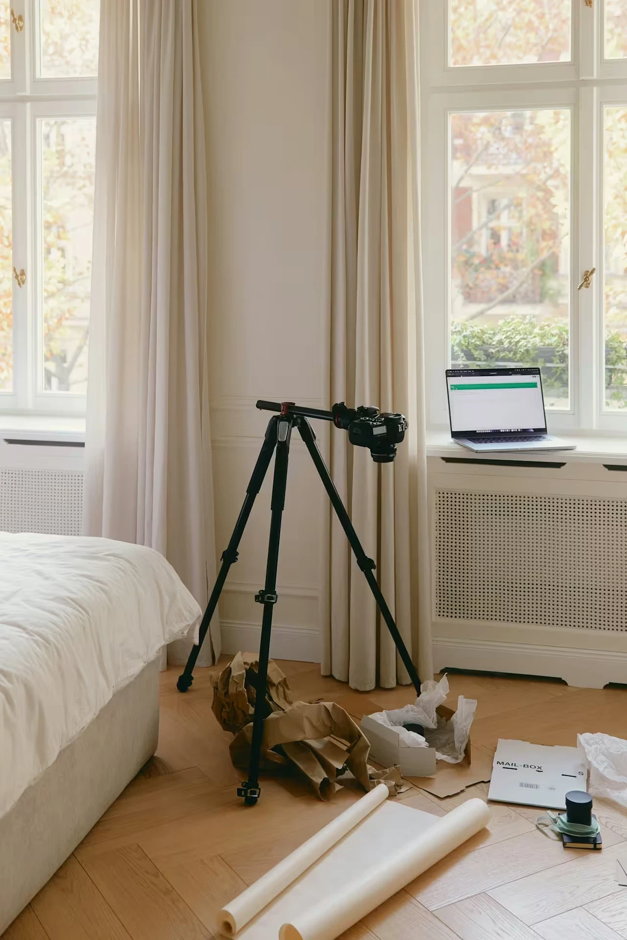 A camera tripod and open laptop sit in a bright bedroom near large curtained windows.