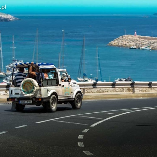 A white open-top vehicle with passengers drives along a coastal road with boats docked in the marina and blue water in the background.