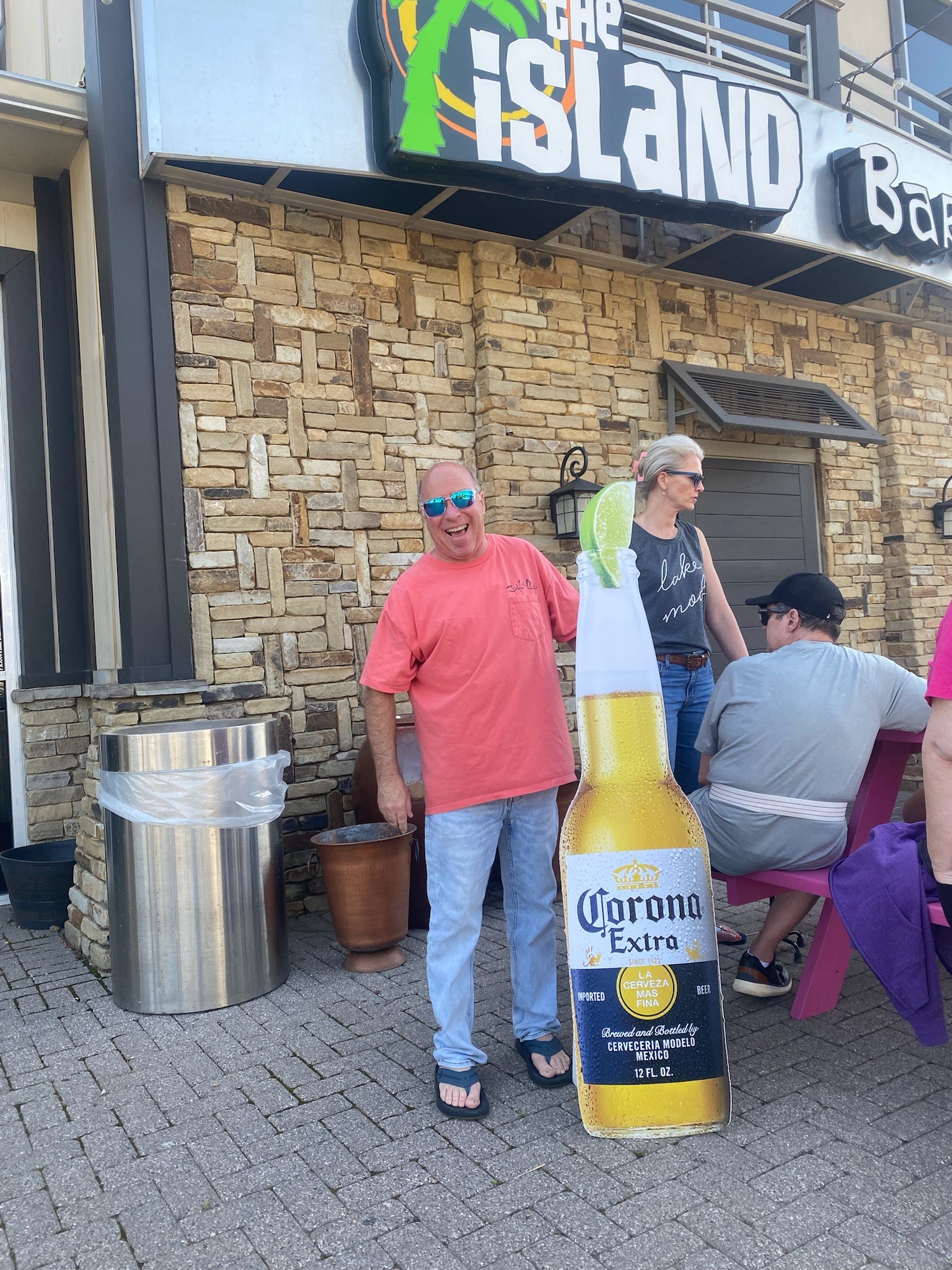 A man in a red shirt stands outdoors beside a life-sized cutout of a Corona Extra beer bottle with a lime, in front of a restaurant with a rustic stone facade.
