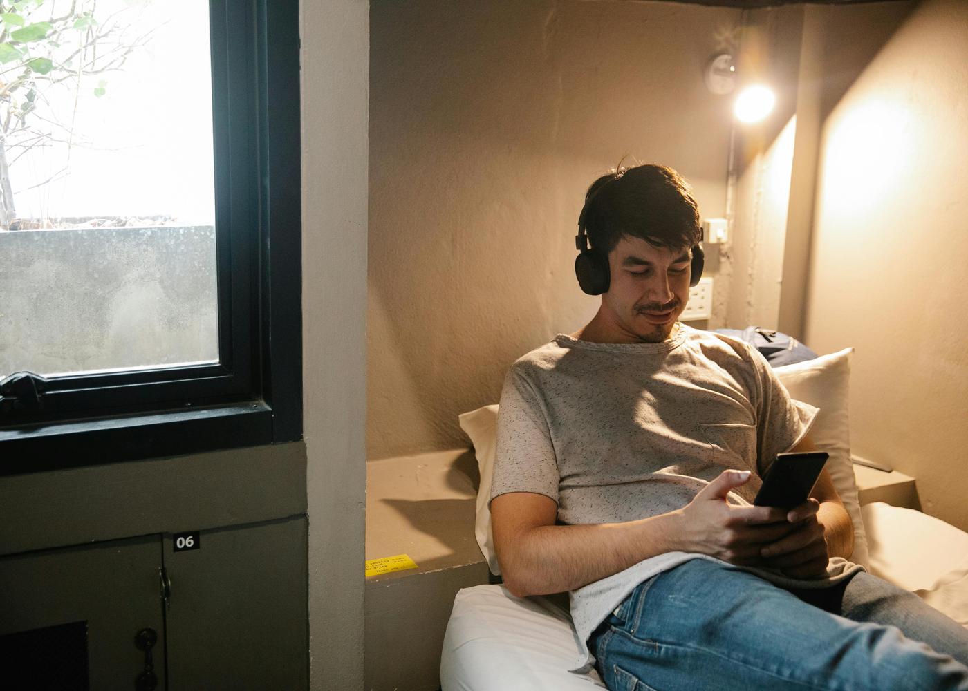 Man with big headphones on sitting upright on a bed looking at his phone