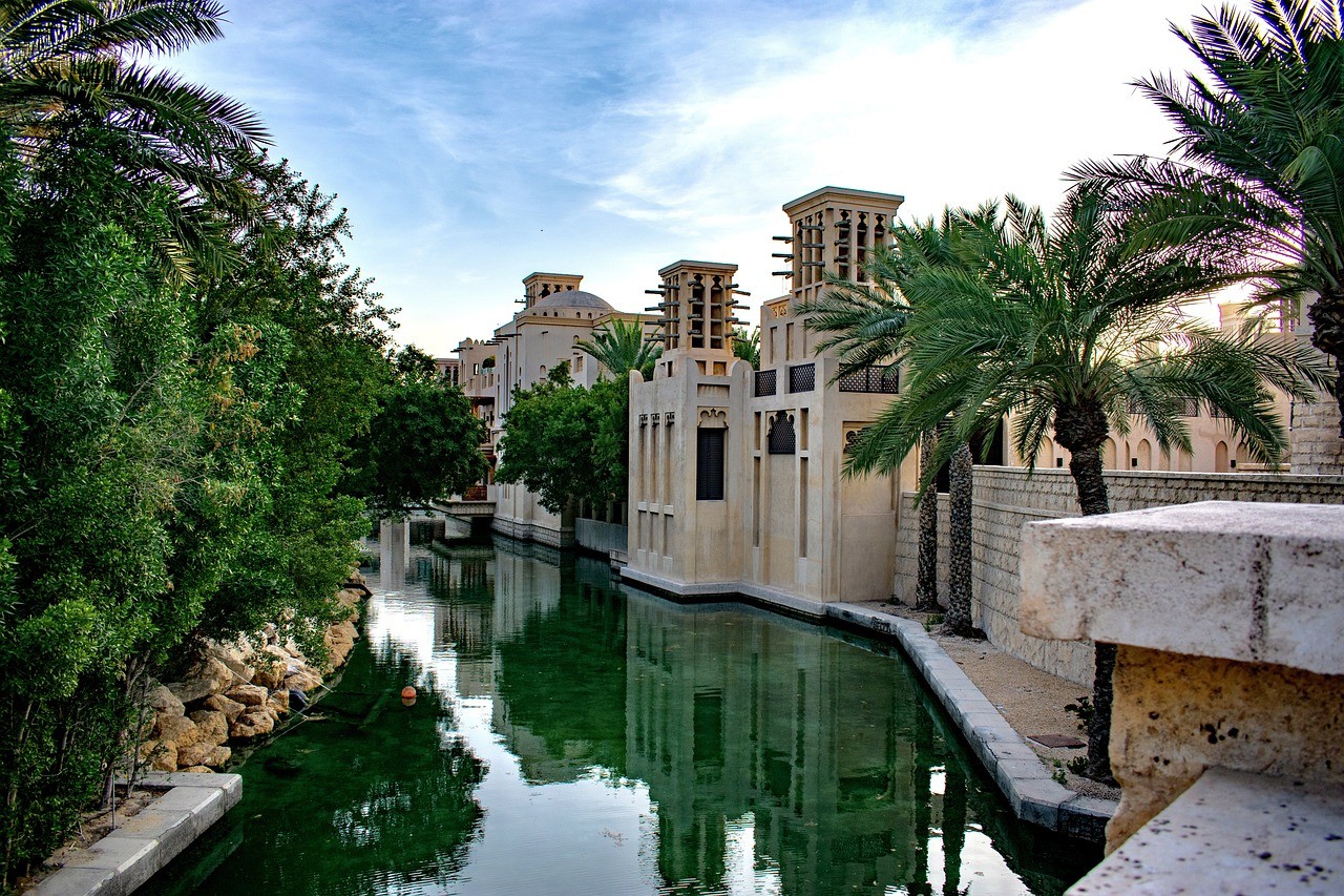 A waterway with traditional beige buildings, palm trees, and calm water reflections.&nbsp;