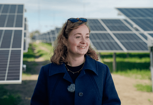 Woman with blonde hair standing and smiling in front of a field of solar panels.