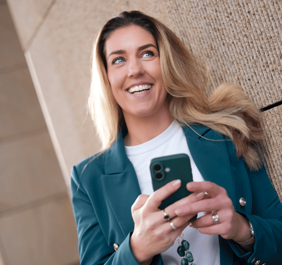 A woman in a teal dress sits in a dimly lit room, focused on her smartphone. A laptop and notepad are on the glass table, suggesting a working environment.