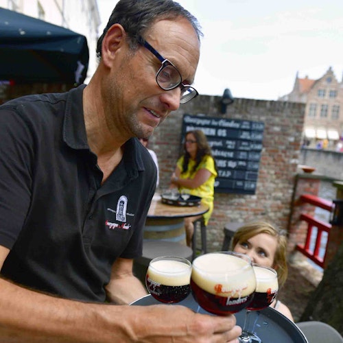 Egy pincér szemüveggel három pohár sört szolgál fel tálcán a vendégeknek egy kültéri kávézóban, ahol a háttérben egy téglafal látható.