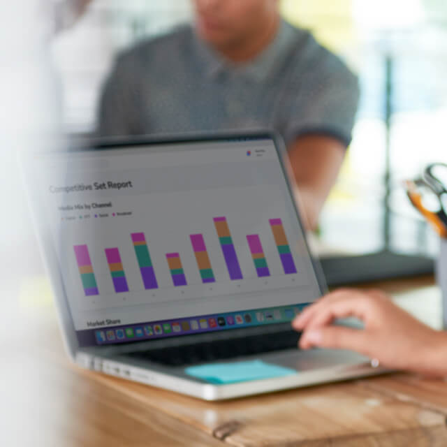 A person uses a laptop displaying a colorful bar chart labeled "Competitive Set Report" at a desk, with blurred people in the background.