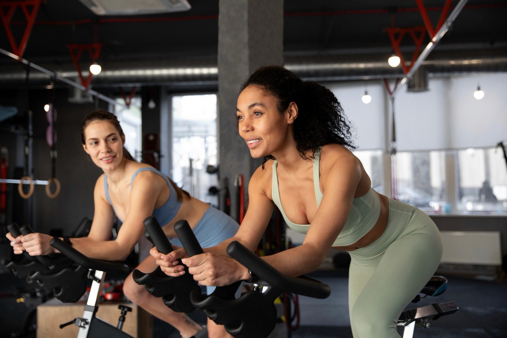 Two women in athletic wear are smiling while riding stationary bikes during an indoor cycling class at a gym.