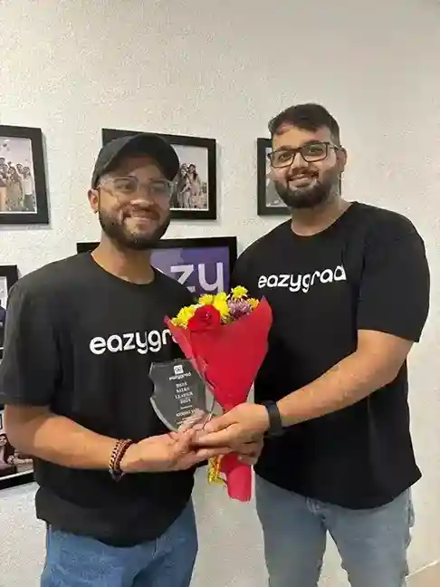 Two people wearing black "eazygrad" t-shirts stand side by side, smiling and holding a flower bouquet and a transparent award, with framed photos on the white wall in the background, representing success in UG/PG online degree programs and unbiased counselling.
