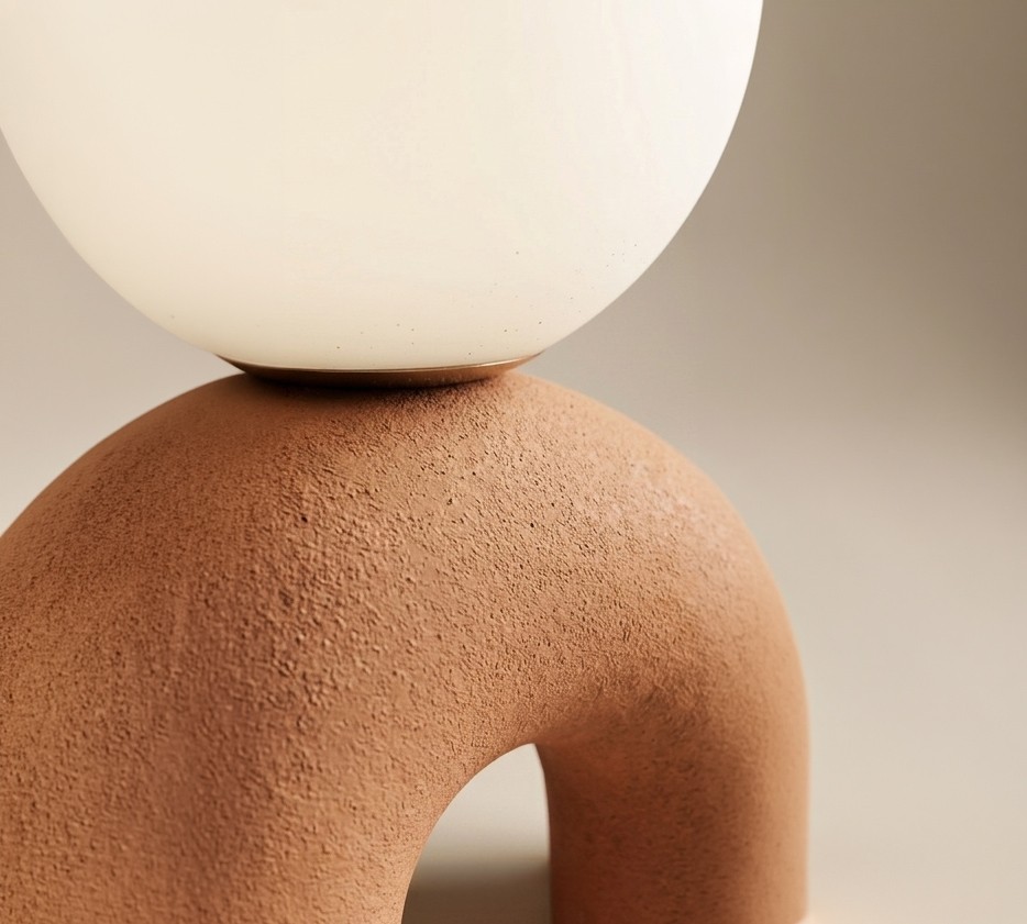 Close-up of a minimalist lamp with a matte terracotta arch base and a frosted white glass globe resting on top, showing the textured surface detail against a soft beige background