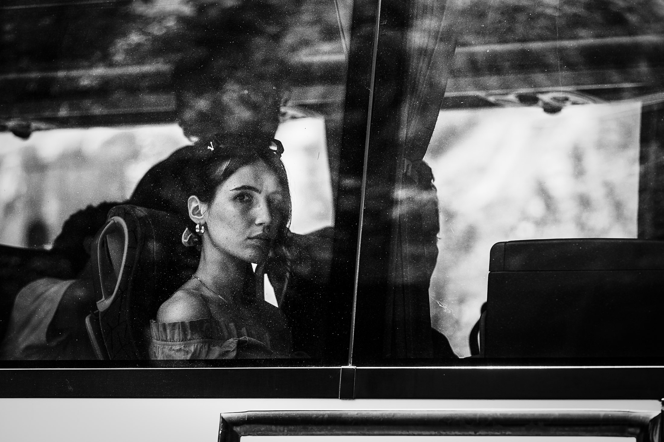 A woman wearing earrings gazes out from inside a bus, her face framed by reflections on the window.