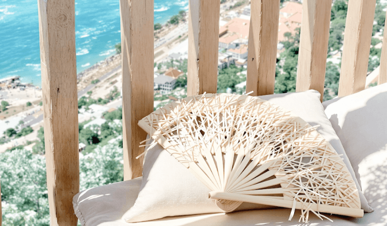 Decorative hand fan resting on cushions with a scenic coastal view below.