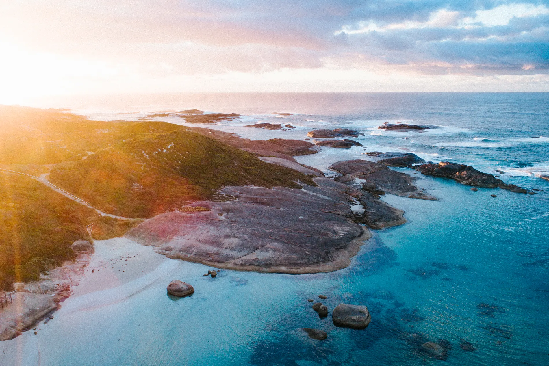 Aerial at low sun over a rocky coastline, pale sand and shallow blue water