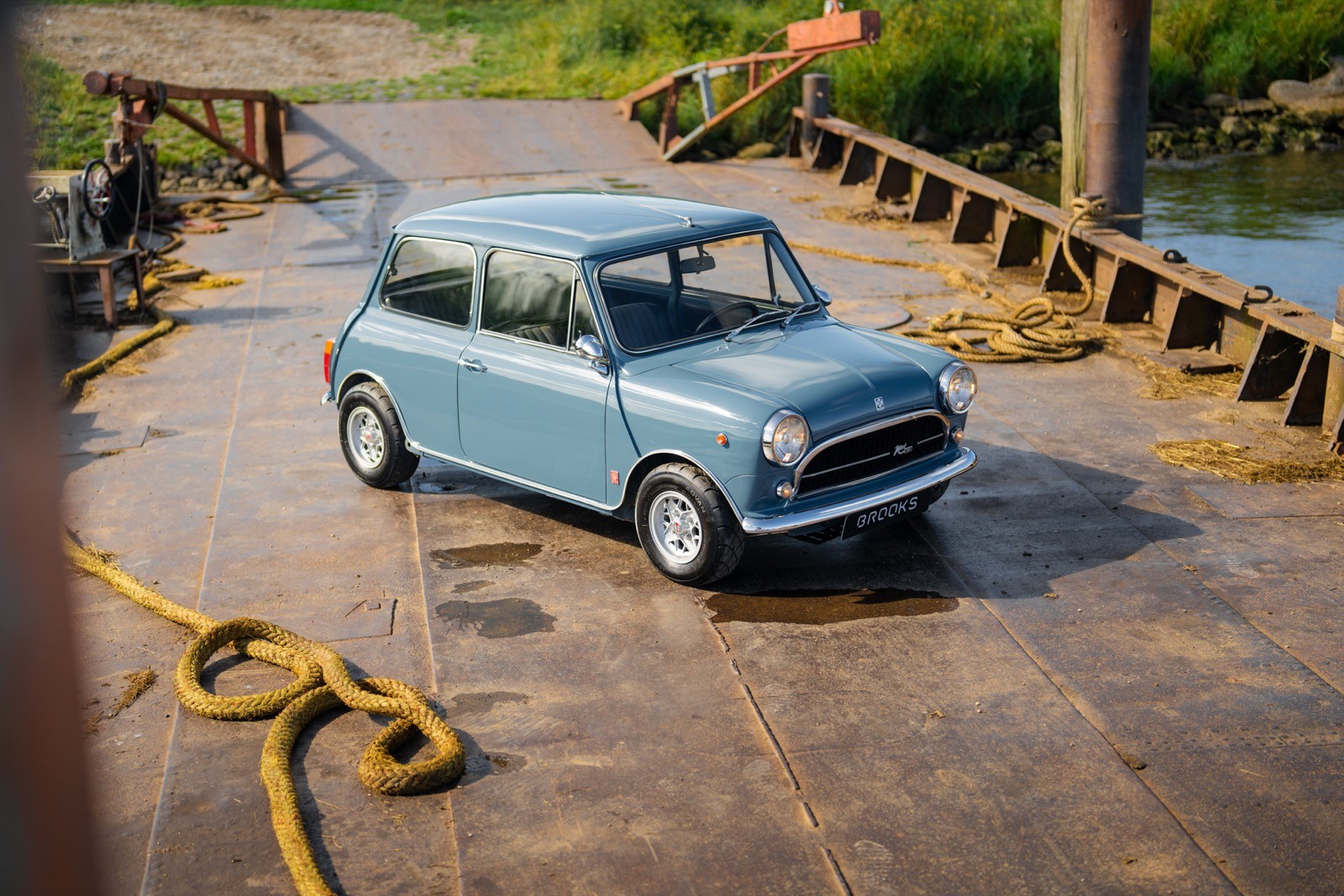 A classic blue car parked on a rustic dock near a body of water, surrounded by greenery and wooden structures.