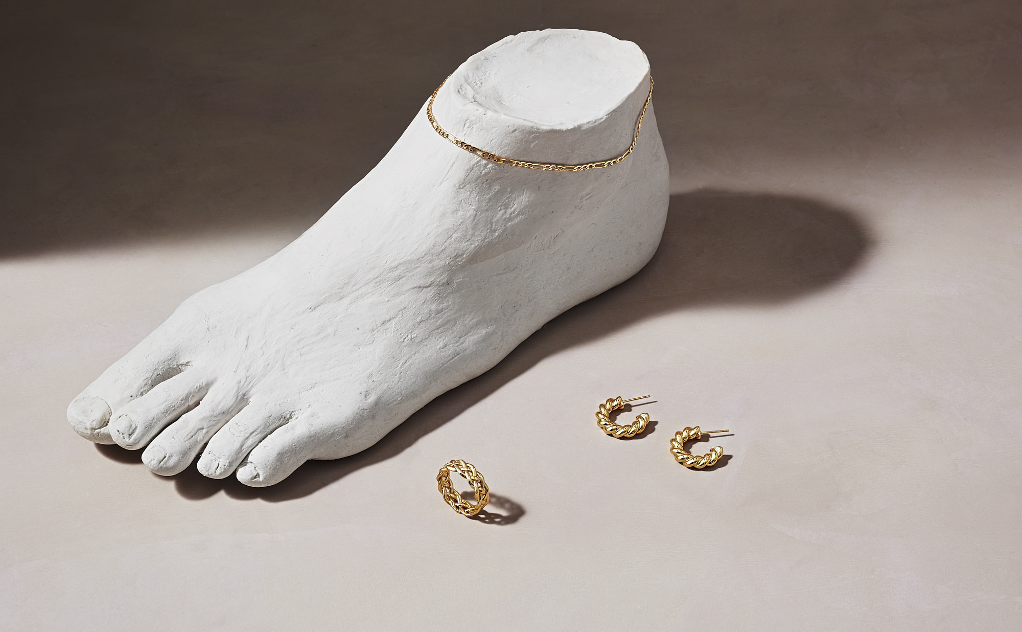 Plaster foot sculpture with gold chain anklet and gold hoop earrings on beige background.