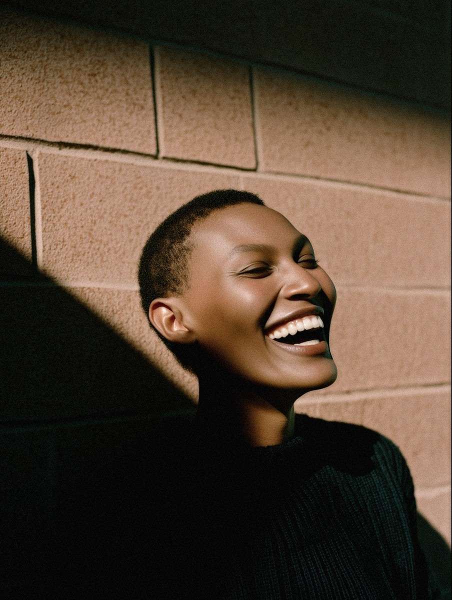 Woman with short hair laughing against a sunlit brick wall.