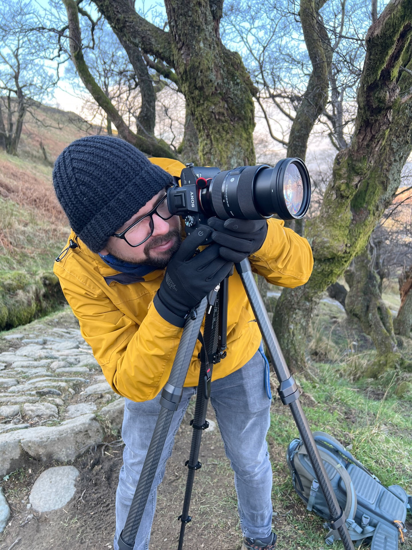 Martin is seen peering through the viewfinder of his camera on a tripod. He's wearing a black wooly hat and gloves.