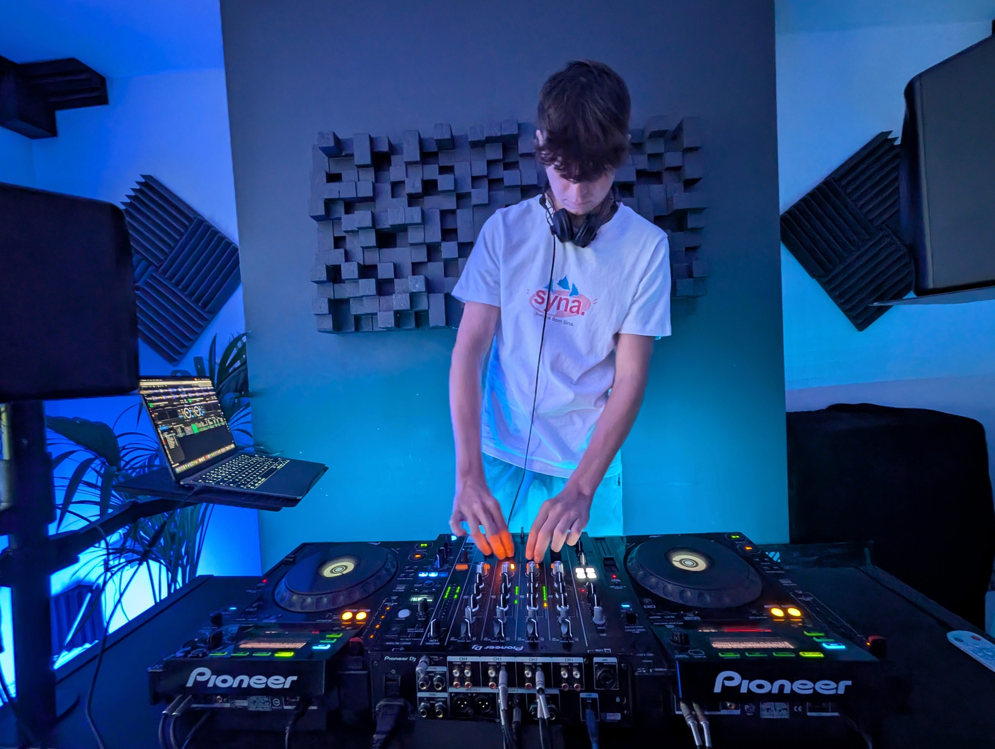 A DJ standing at a mixing board, wearing headphones and a white t-shirt, working with Pioneer DJ equipment.