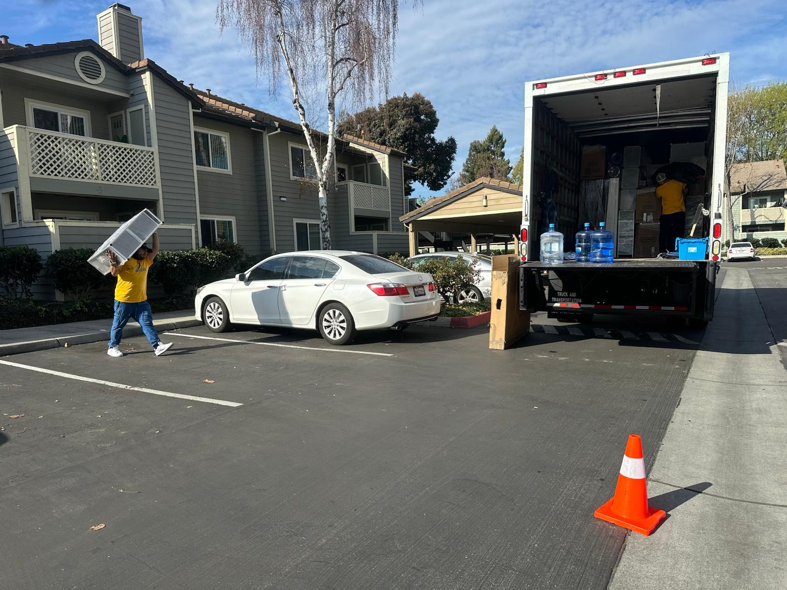 Zapt Movers Team at Apartment Complex Two Zapt Movers team members in yellow shirts are transporting boxes and furniture from an apartment complex into a moving truck parked by the curb. Packing materials and large water jugs are visible inside the truck. An orange safety cone marks the loading area for safety.