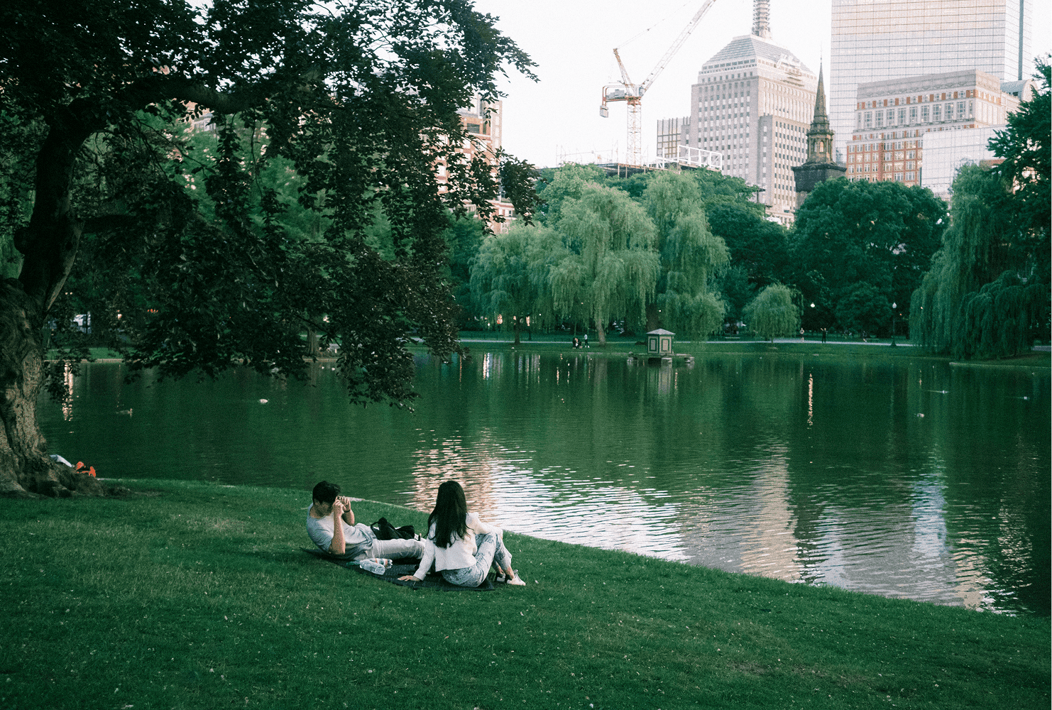 a couple laying on the grass next to the lake in central park. The man is taking a photo of the woman. 
