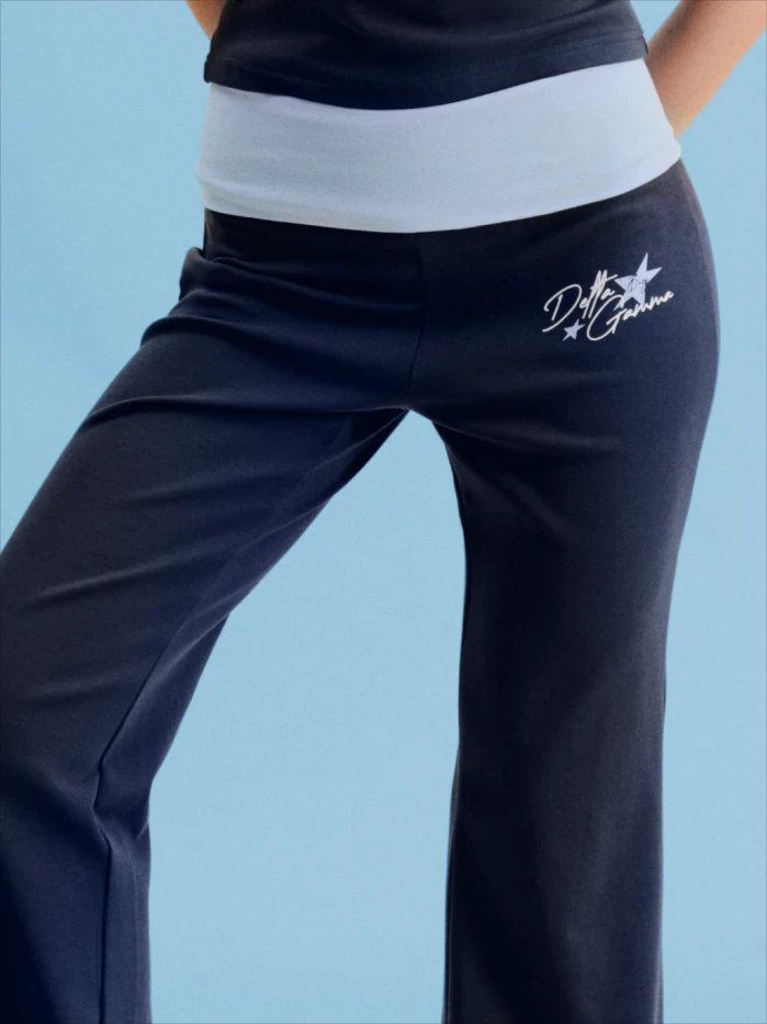 Close-up of a woman wearing navy fold-over waistband flare sweatpants with a white star and "Delta Gamma" script logo embroidered on the hip, set against a light blue background.