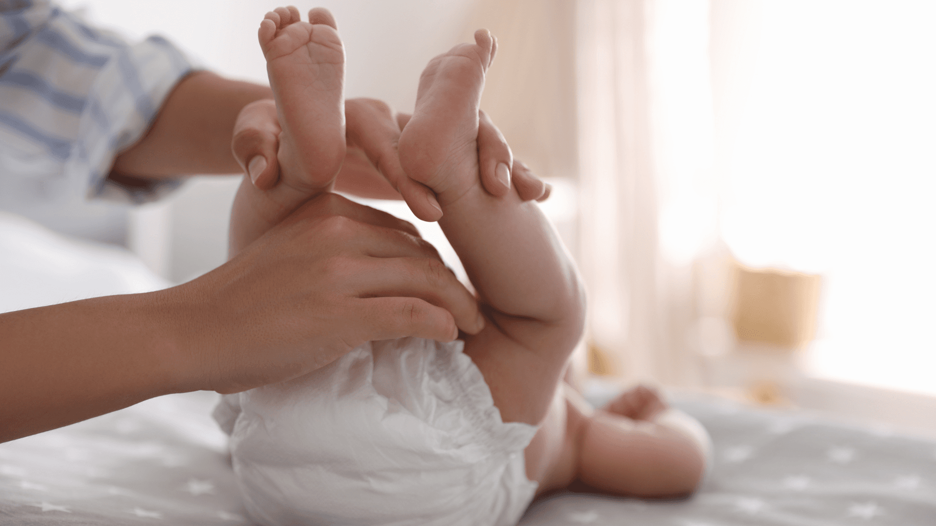 Alt tag: Caregiver gently holding a baby’s legs up while changing a diaper on a soft surface in a bright, cozy room