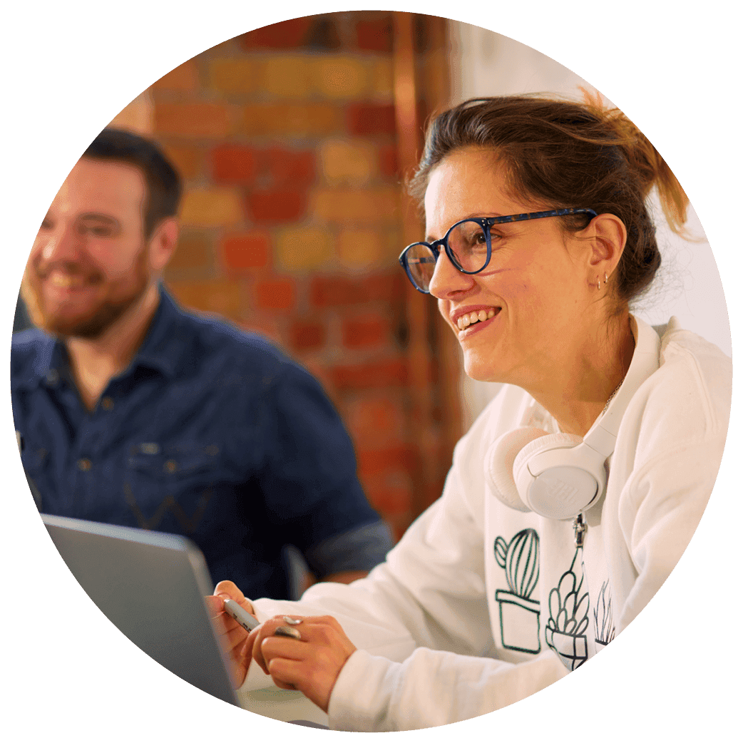 Two people seated at a table working on laptops, with one wearing headphones around their neck.