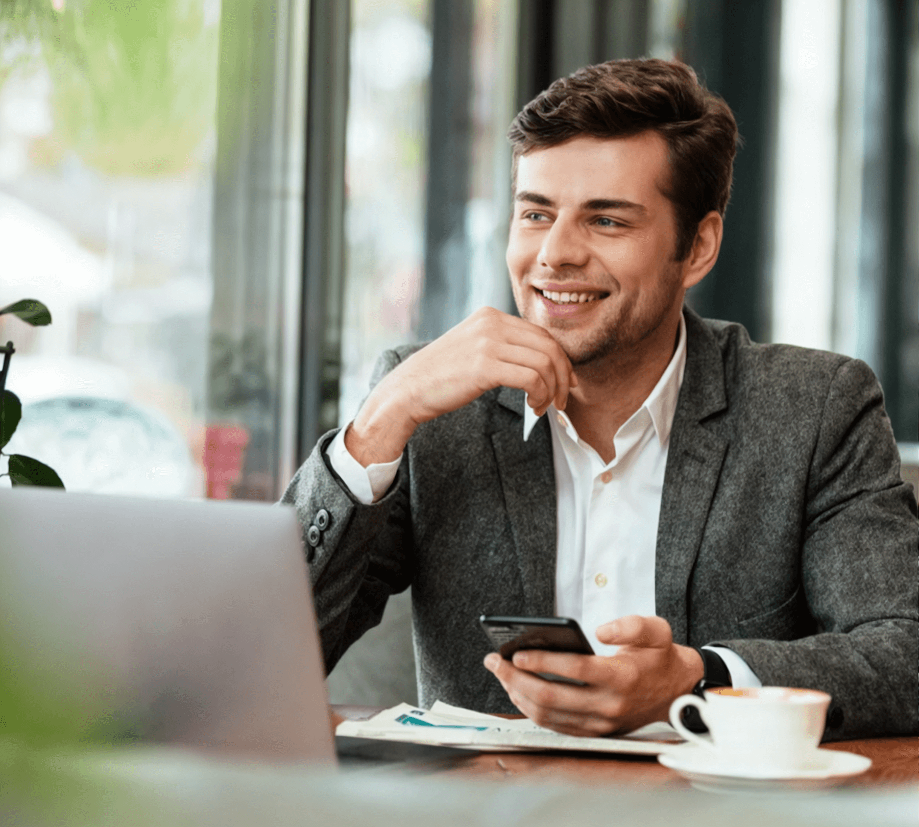 A man in a suit sits at a table with a laptop and a cup of coffee. He is smiling and looking away from the camera.