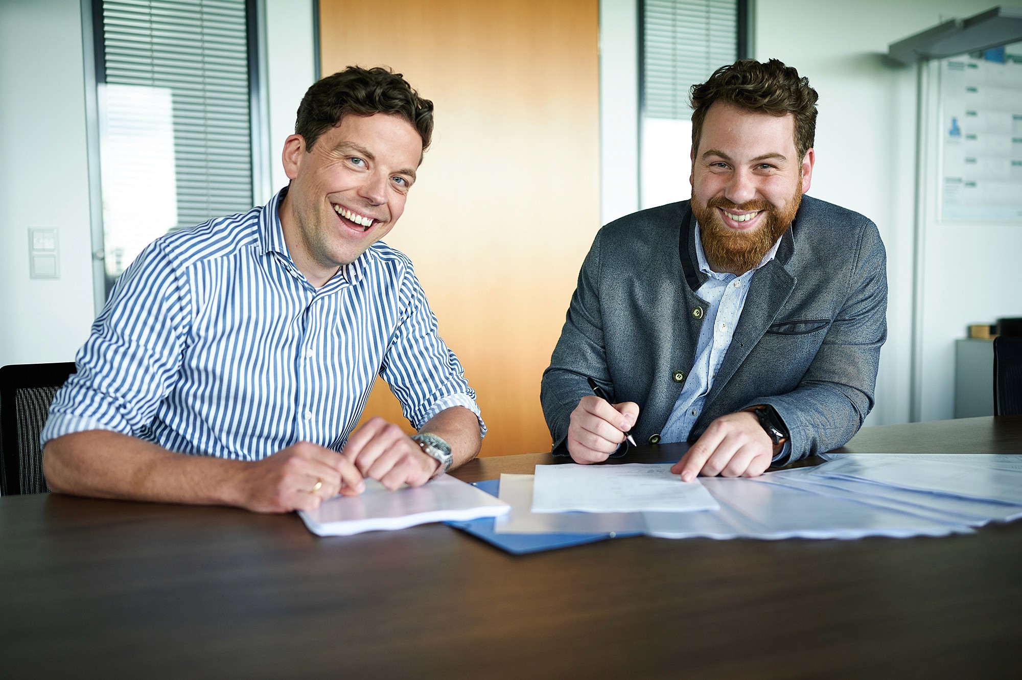 Two men sitting at a table with documents spread out, both laughing at the camera.