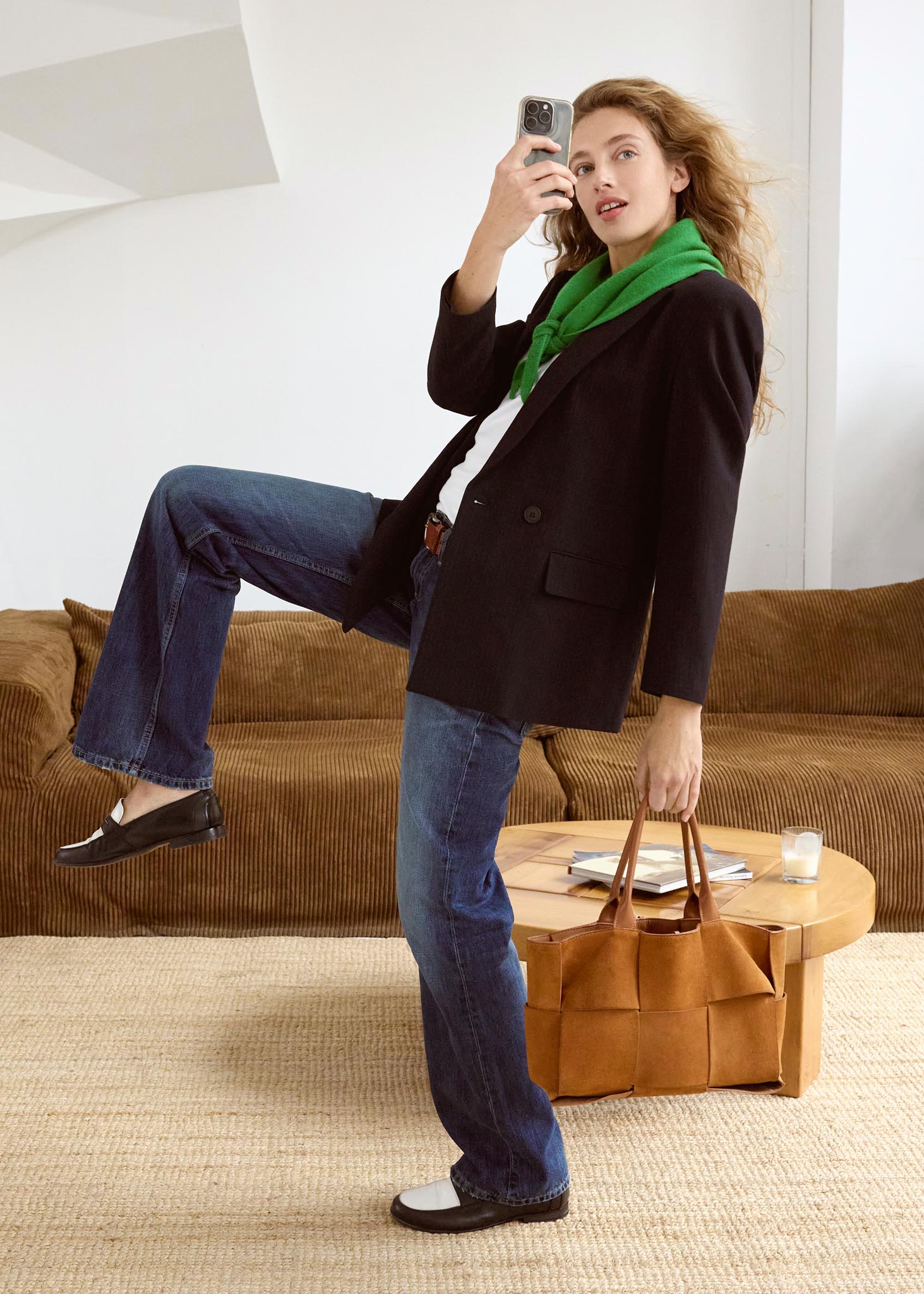 Femme debout dans un salon, prenant une photo avec son téléphone, portant une veste sombre et un foulard vert