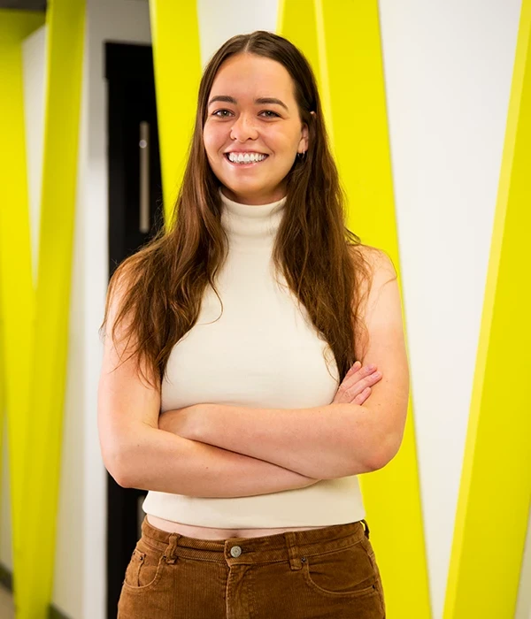 A person stands with arms crossed in a brightly lit hallway featuring bold yellow structural elements. Casual clothing and strong lines give the image a confident, contemporary feel.