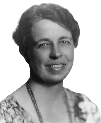 Black and white portrait of a woman with short, neatly styled hair, smiling warmly at the camera. She wears a floral-patterned dress and a simple necklace, exuding a sense of grace and kindness. The soft lighting and gentle expression convey approachability and warmth.