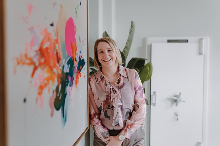 Geschäftsführerin in gemusterter Bluse steht neben einem bunten Kunstwerk an der Wand und spricht mit einer Person außerhalb des Bildes.