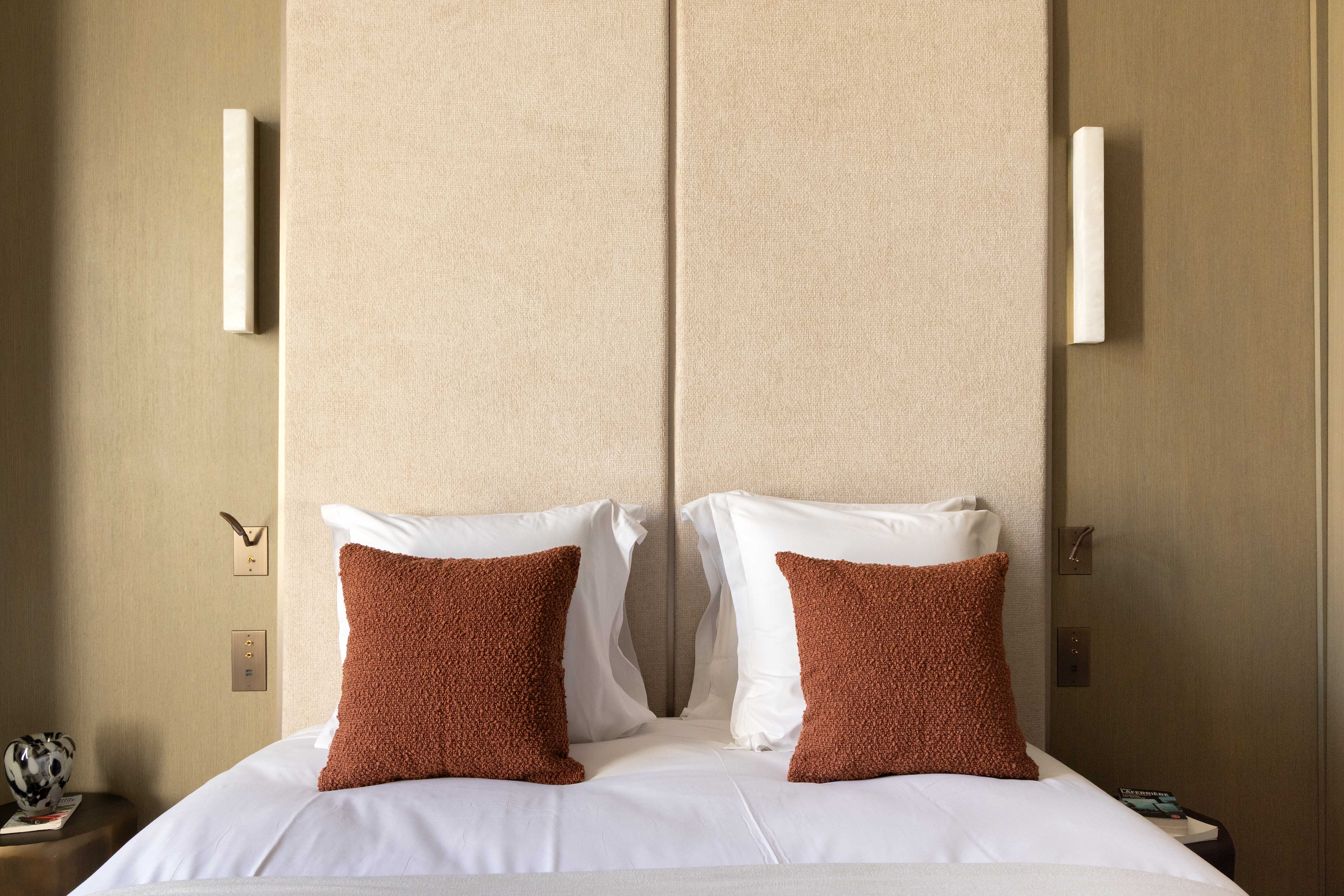 Detail de la tete de lit velours beige avec coussins terracotta, appliques murales et interrupteurs dores
