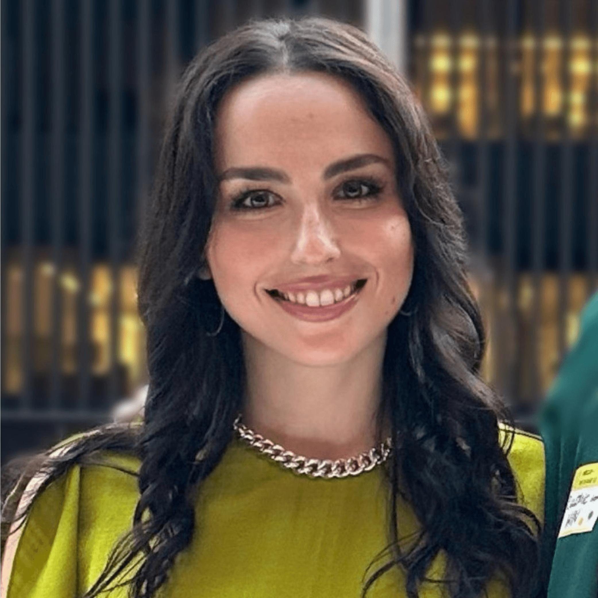 A young woman with dark hair smiles in a casual outfit, standing in a modern indoor setting.