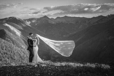 Black-and-white portrait of the couple kissing as a long veil blows across the mountain view.