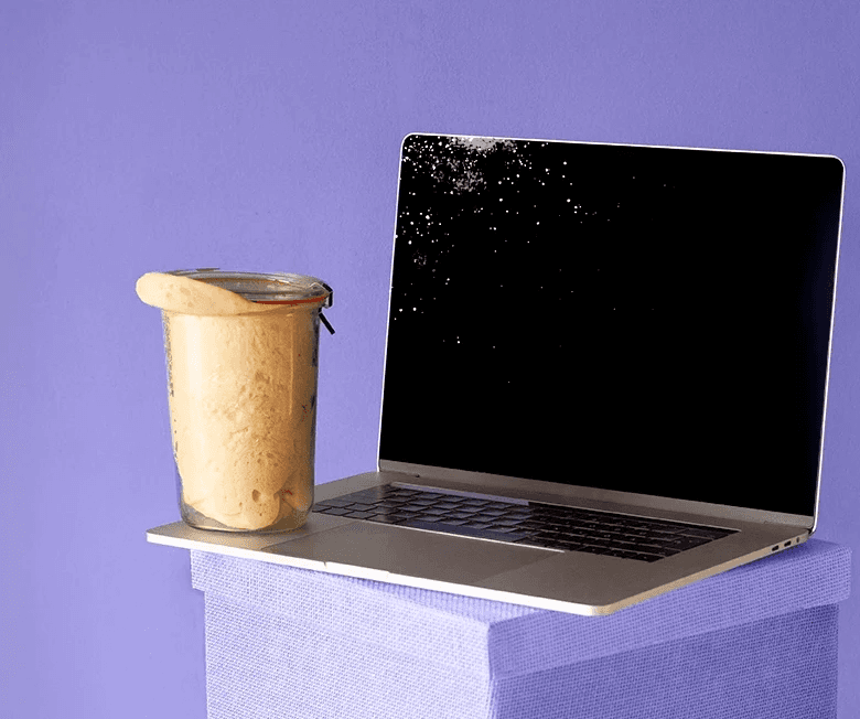 Laptop placed on a lila box, next to a jar with sourdough almost exiting it, on a purple background