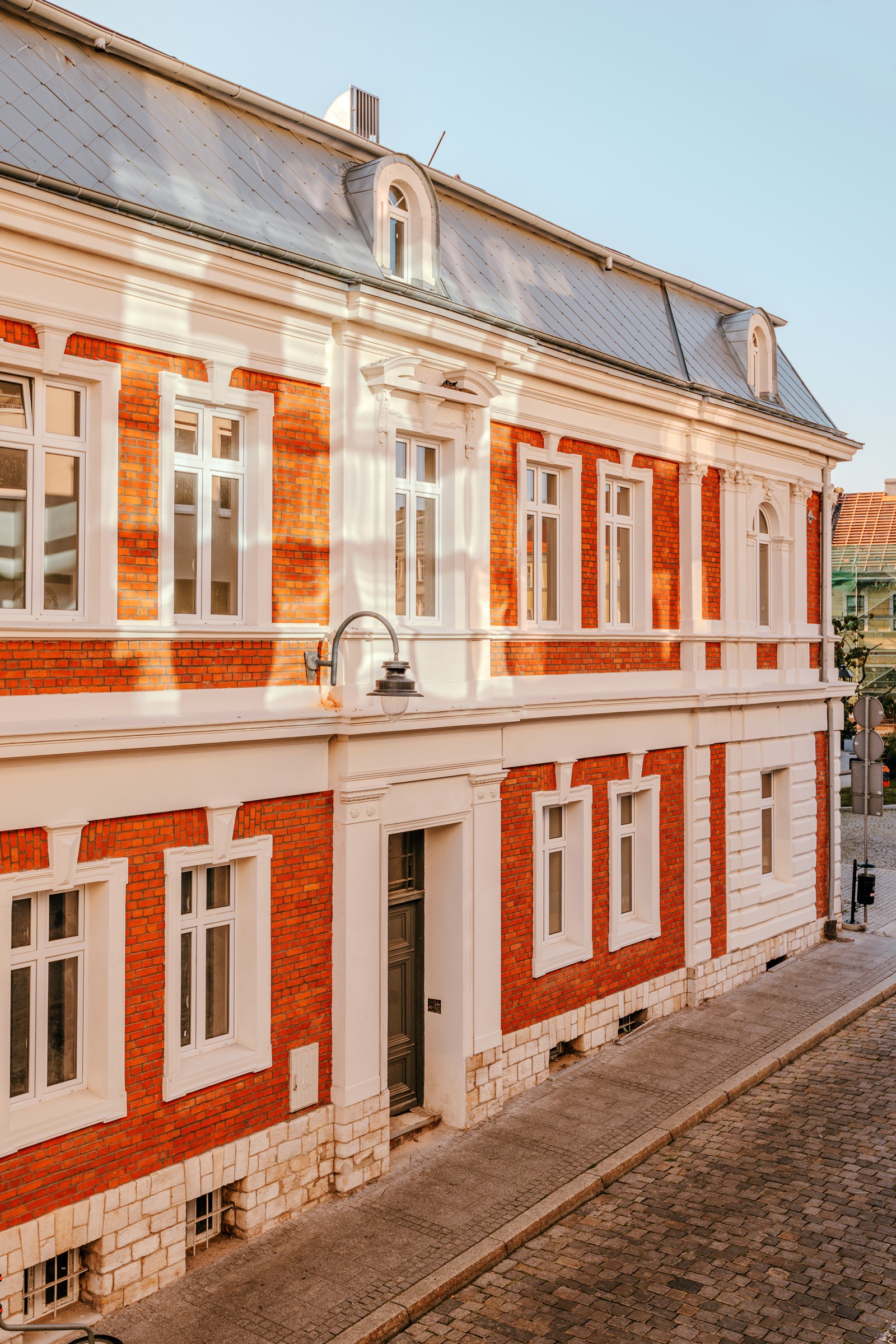 Piękny budynek w stylu klasycystycznym z czerwonymi cegłami. Na rogu ulicy z białymi elementami. Stoliki i parasole na zewnątrz.