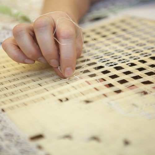 Close-up of a hand weaving thin thread through an off-white, grid-patterned fabric.