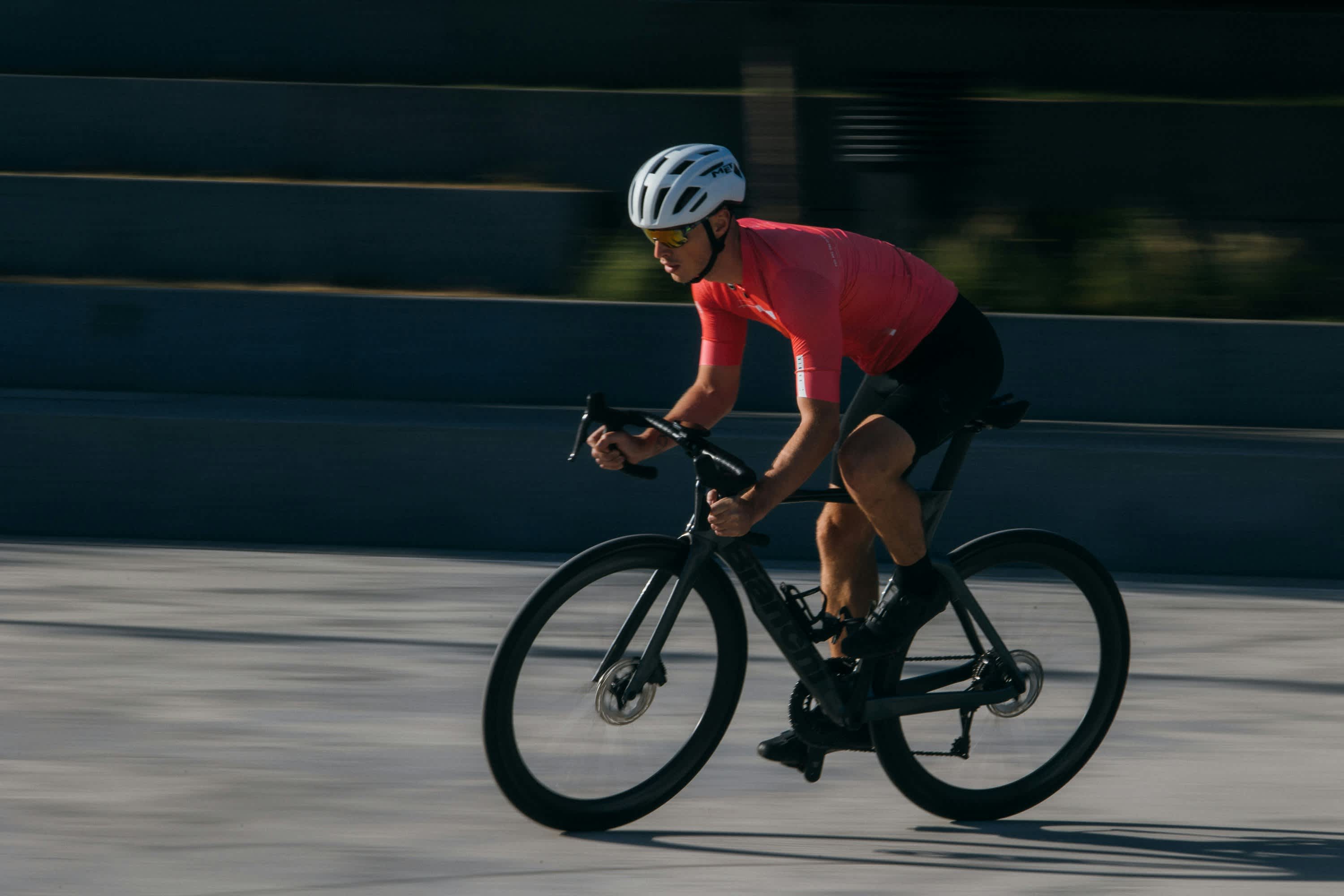 Road cyclist wearing a red jersey and white helmet riding fast on a paved track with motion blur in the background.