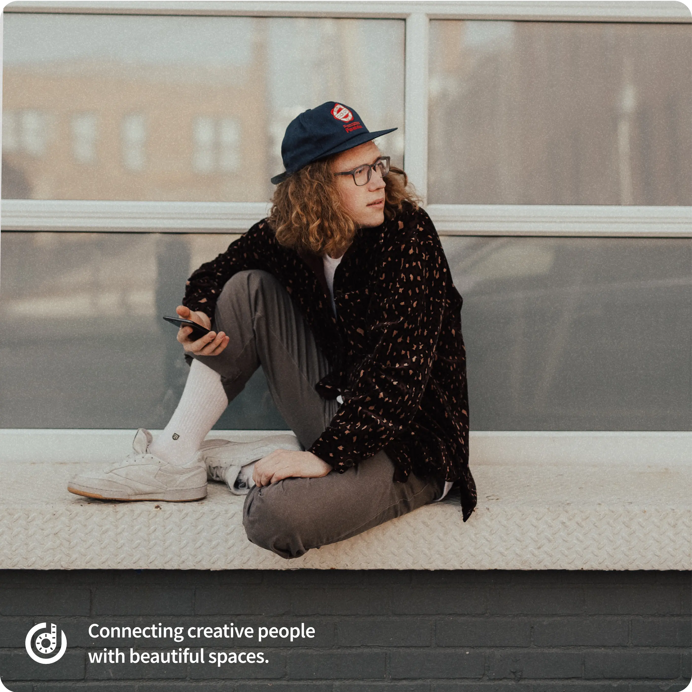 A young person with curly hair wearing glasses, a blue cap, a patterned jacket, grey pants, and white sneakers sits casually on an outdoor ledge while holding a smartphone. In the bottom left corner, there's a logo featuring a circular design with a red film reel forming the letter 'd' and the text 'Connecting creative people with beautiful spaces.'