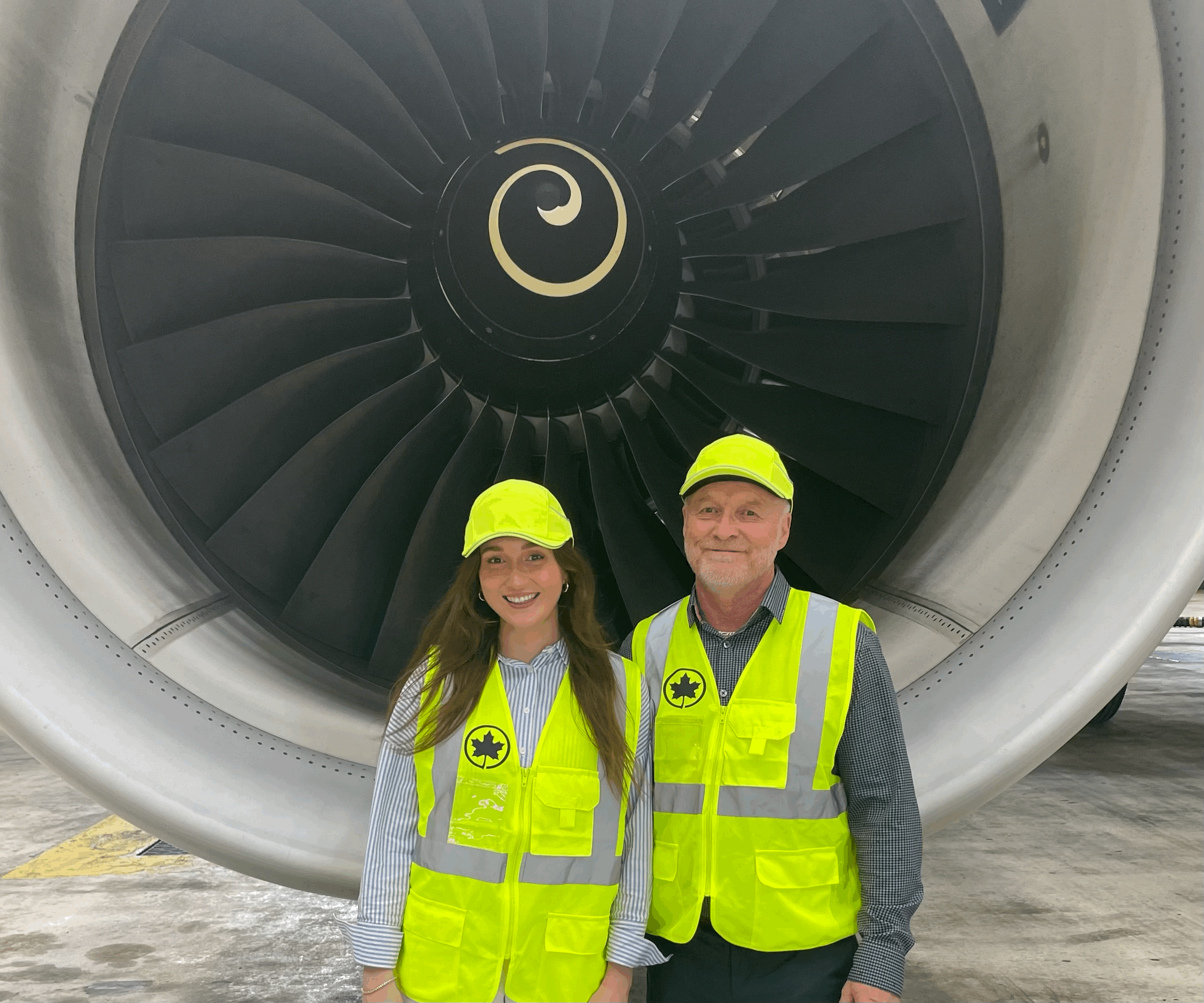 Deux personnes portant des vestes et casquettes de sécurité jaune fluorescent se tiennent devant un immense moteur d’avion dans un hangar, souriant face à la caméra.
