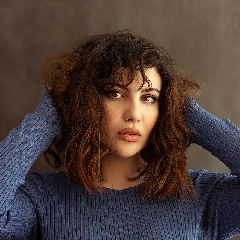 A woman with wavy, shoulder-length brown hair and bangs wearing a blue ribbed sweater, posing with her hands behind her head against a dark studio background.