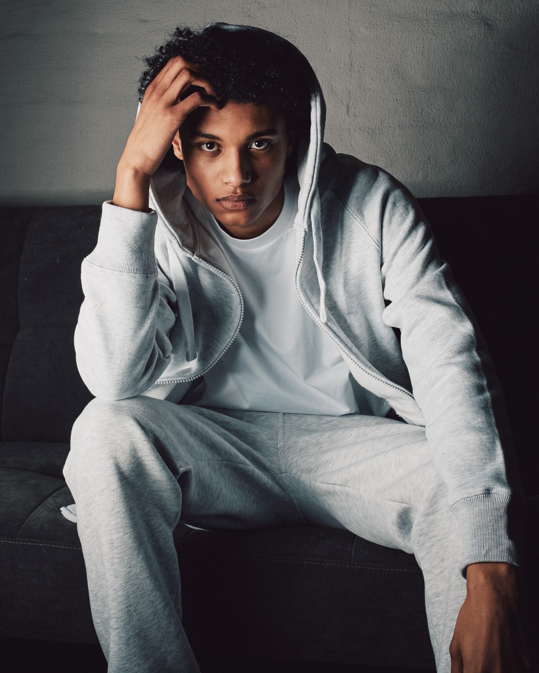Person dressed in ligh grey zip hoodie and sweatpants, sitting on a couch in a dark, contrastful setting