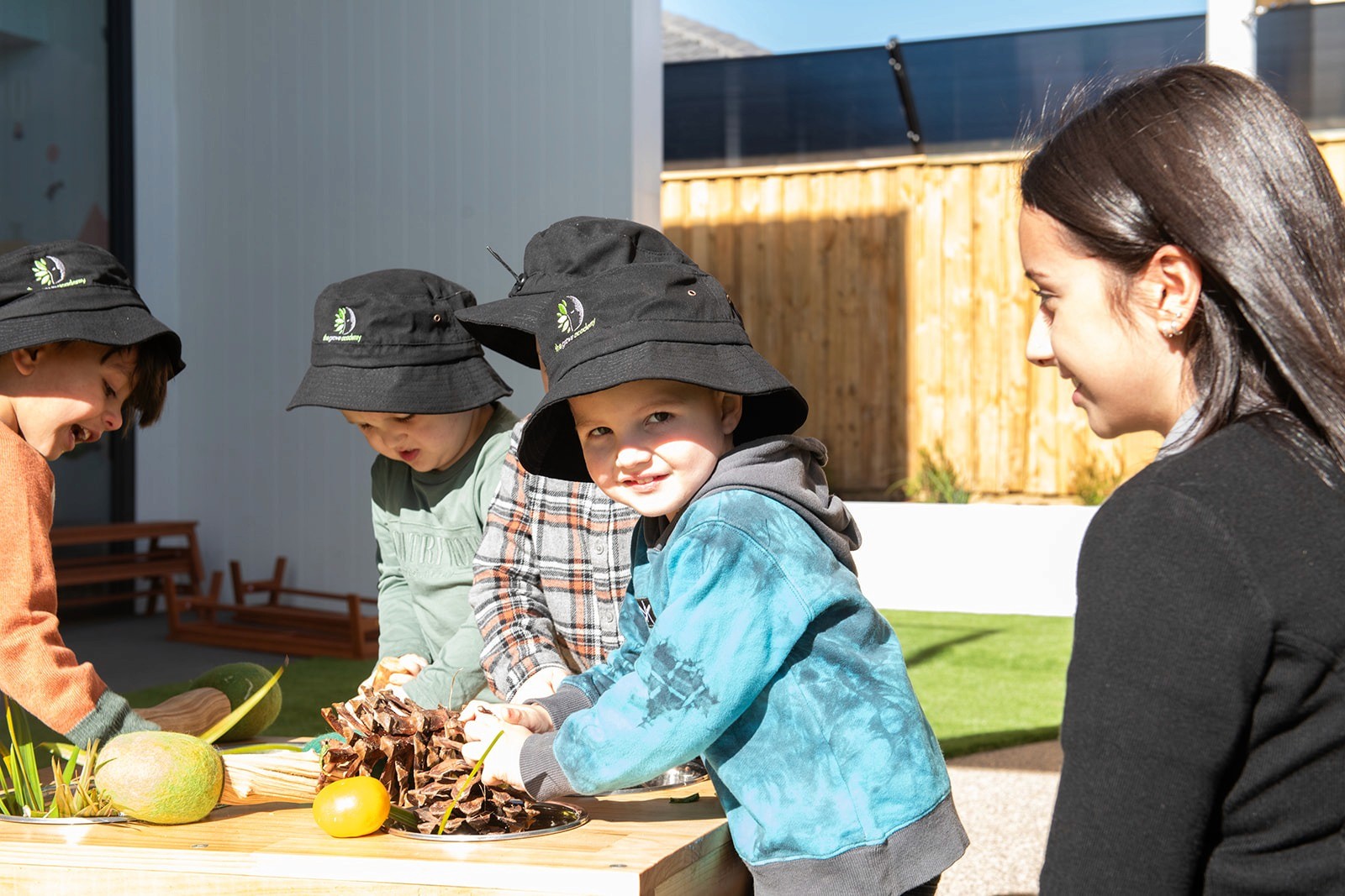 Smiling young woman with long hair standing against a dark green background, holding a finger to her chin.
